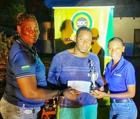 Ashley Simms (right), Sandals regional public relations coordinator for Montego Bay Region, assists Evett Malcolm, Social Development Commission Field Supervisor in handing over The Sandals Trophy to captain Tiray Thoston at the presentation ceremony for the Retirement Community Football League recently.