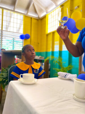 Grade 4 student at Negril Primary School, De-raina Kara Gordon watches keenly and Sandals Negril Restaurant Supervisor, Keithia Duncan briefed students on the different glasses placed at the dinner table.