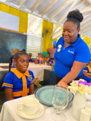 Khari-Grace Andries, student from Negril Primary School being taught on plate placement by Sandals Negril’s Keithia Duncan.